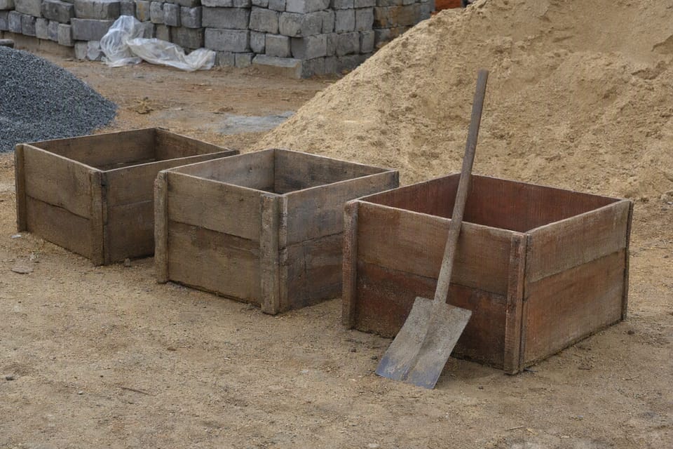 Three empty wooden measuring boxes in a row beside a pile of sand and a shovel