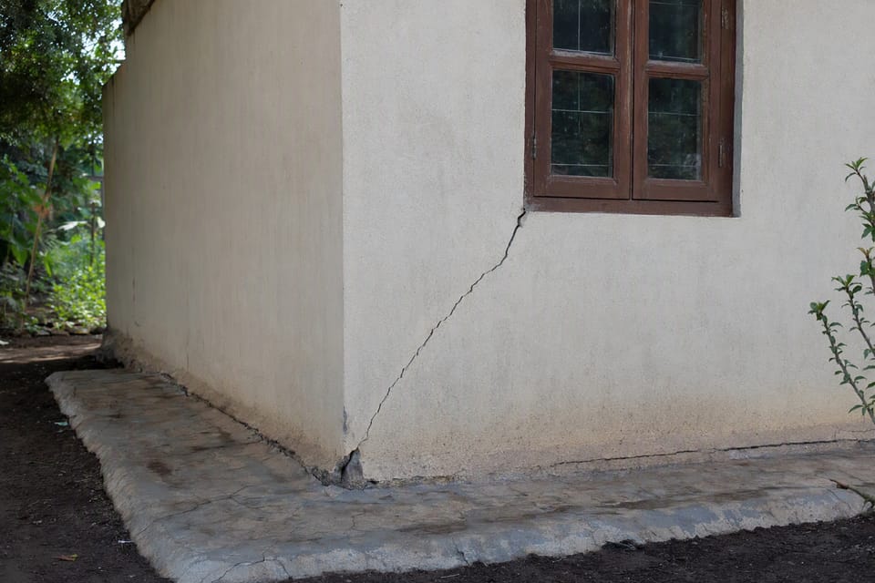 A house corner with a long diagonal crack from the window to the ground and a cracked concrete apron over dark soil