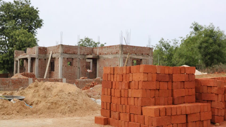 Neat stacks of plain red clay bricks beside a small pile of sand on a building plot with a half-built house behind