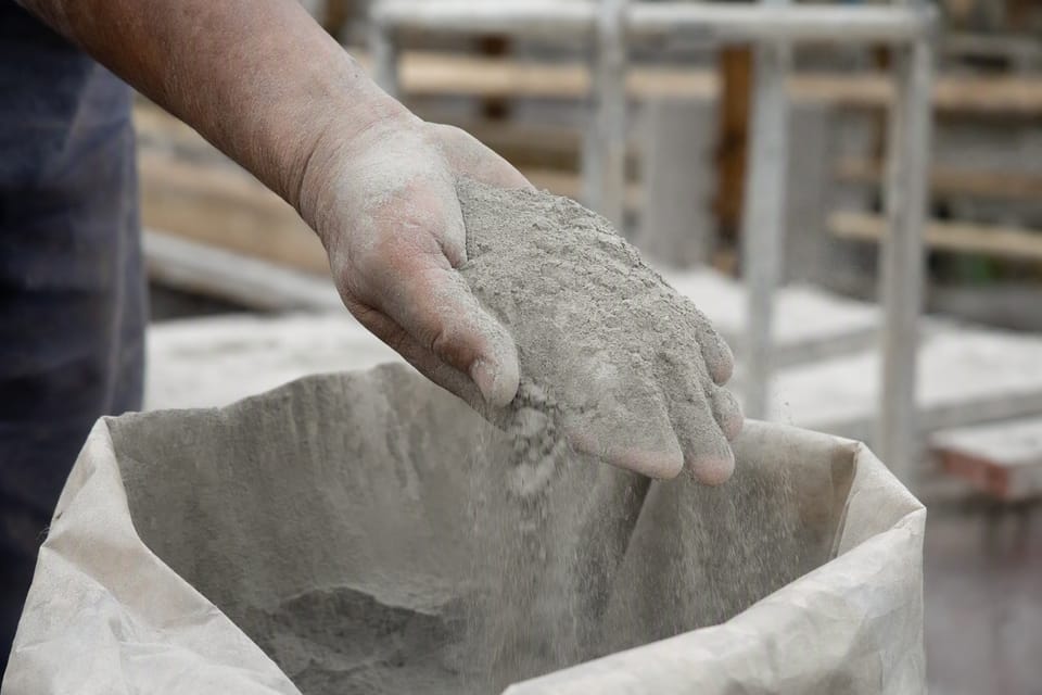 A worker's hand letting fine grey cement powder pour into an open cement bag