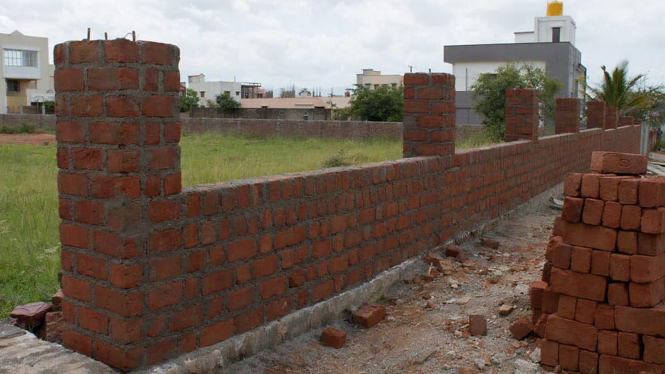 A brick compound wall under construction with square brick pillars at intervals and stacks of bricks beside it