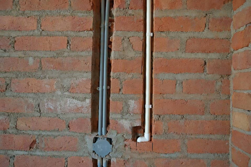Grey electrical conduits and a white water pipe fixed in chases in a brick wall before plastering