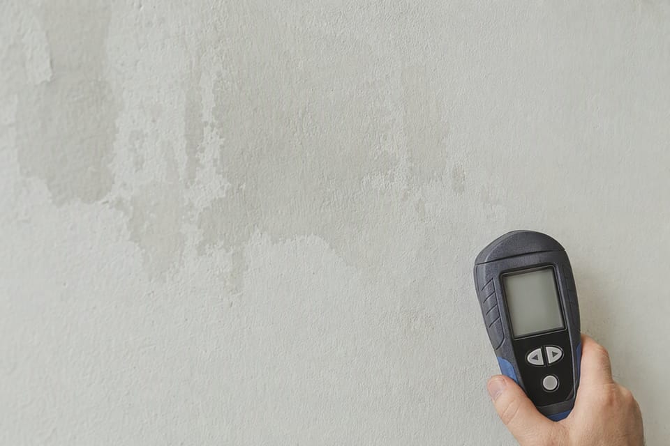 A hand holding a moisture meter against a plastered wall with a faint damp stain