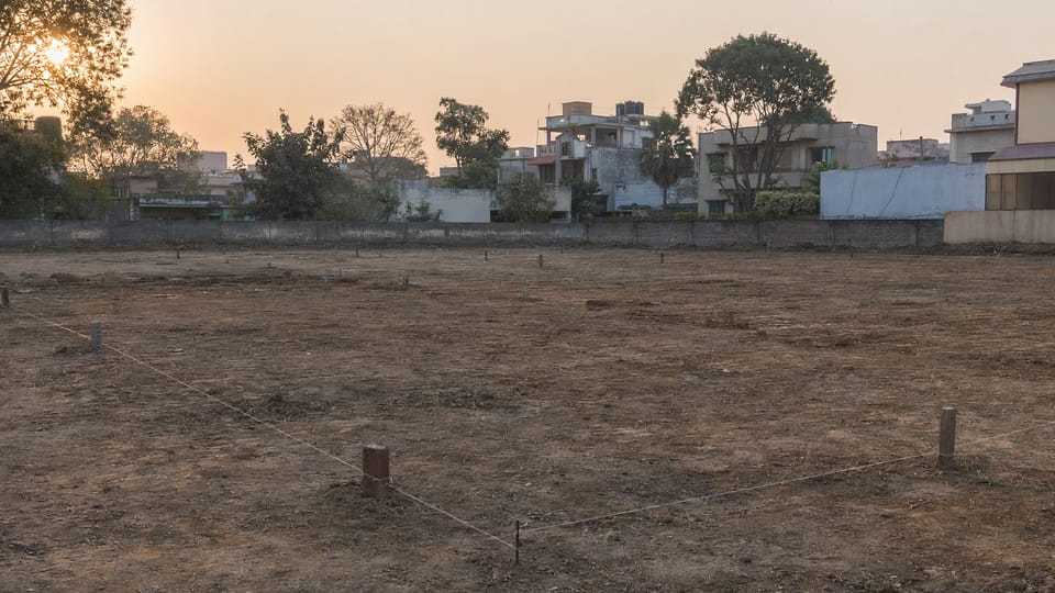 An empty cleared residential plot in a north Indian city at dawn with marker pegs and string lines