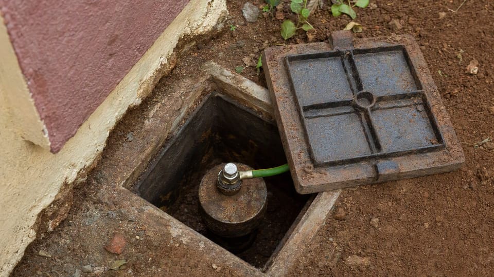 A small earth pit chamber in the ground beside a house wall, its metal cover lifted to show the electrode top with a green-and-yellow wire bolted on