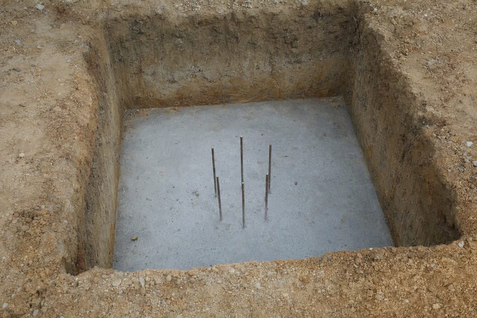A square excavated footing pit with a level grey base and a few short steel bars standing in it