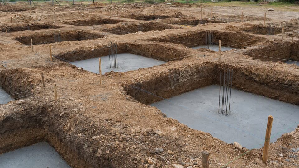 Square excavated footing pits in a grid on a building plot, each with a level grey base and short steel bars