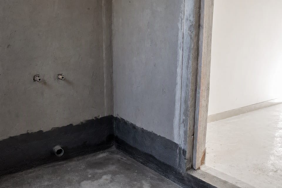 A bathroom with grey cement plaster on the walls and a dark waterproof coat at the bottom, and a white gypsum-plastered room seen through the doorway