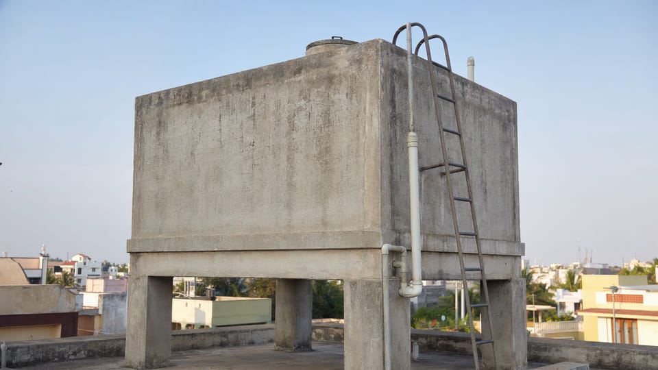 A rectangular concrete overhead water tank on short columns on the terrace of an Indian house with a pipe and a ladder