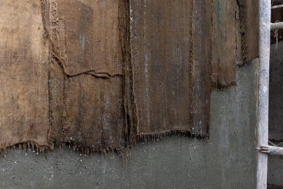 Wet hessian sacking hanging over a freshly plastered grey wall, with water droplets and a scaffold pipe at the side