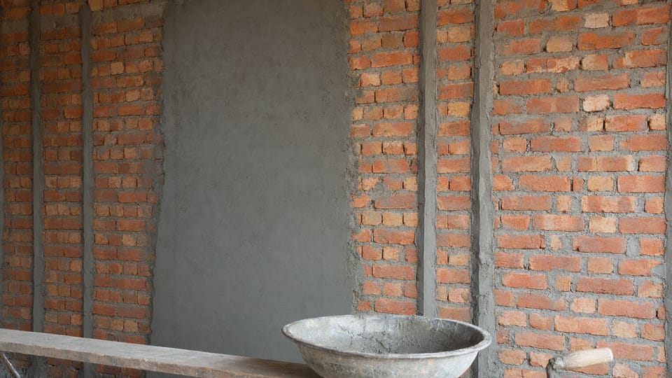 A brick wall with one smooth plastered section, vertical guide strips, a mortar pan and a trowel on a plank