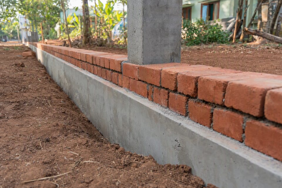 A long grey concrete beam at ground level with courses of red brick laid on it and a square column rising from it