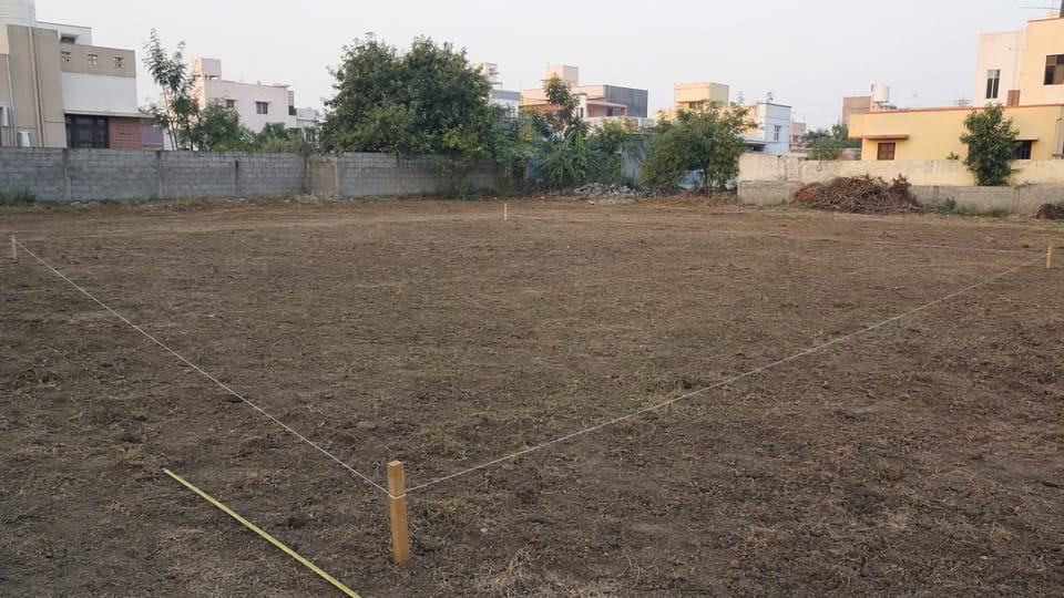 An empty rectangular residential plot with wooden boundary pegs joined by string and a yellow measuring tape along one edge