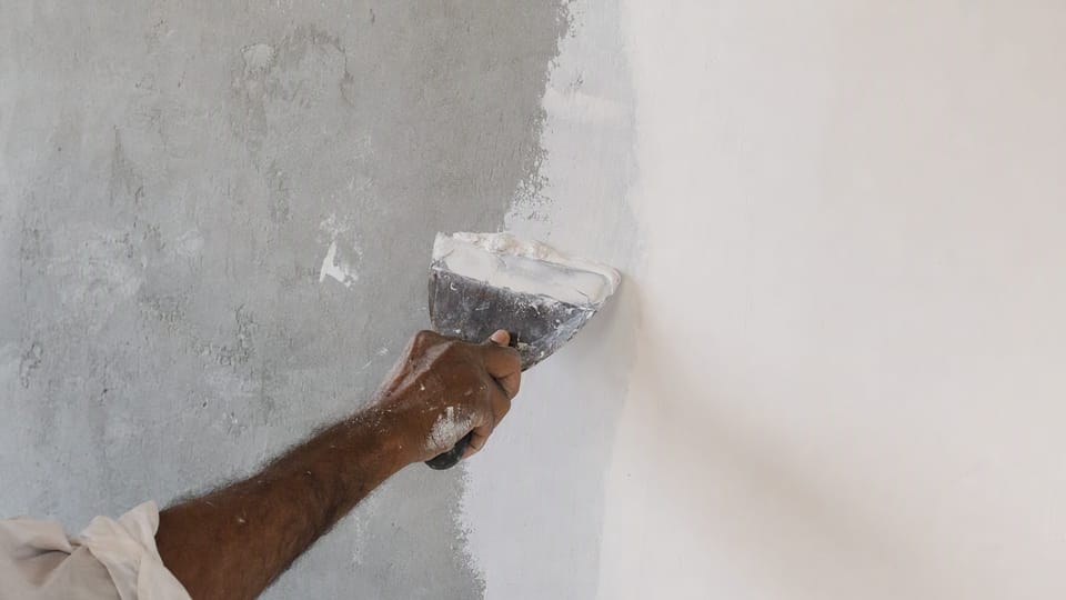 A worker's hand applying white wall putty with a steel blade, half the wall still grey plaster