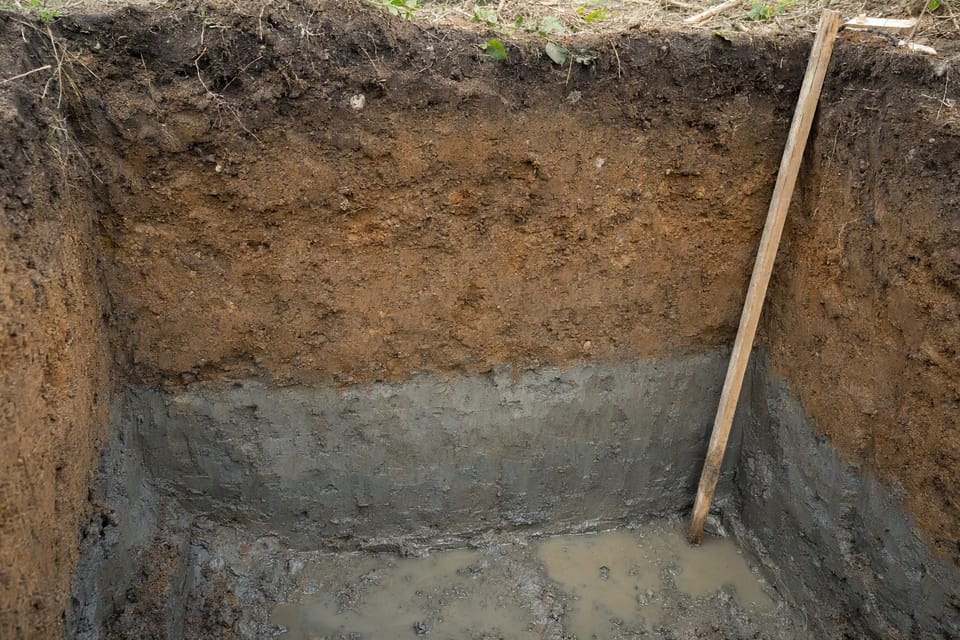 A trial pit wall showing dark topsoil, brown sandy clay and grey clay, with groundwater collecting at the bottom
