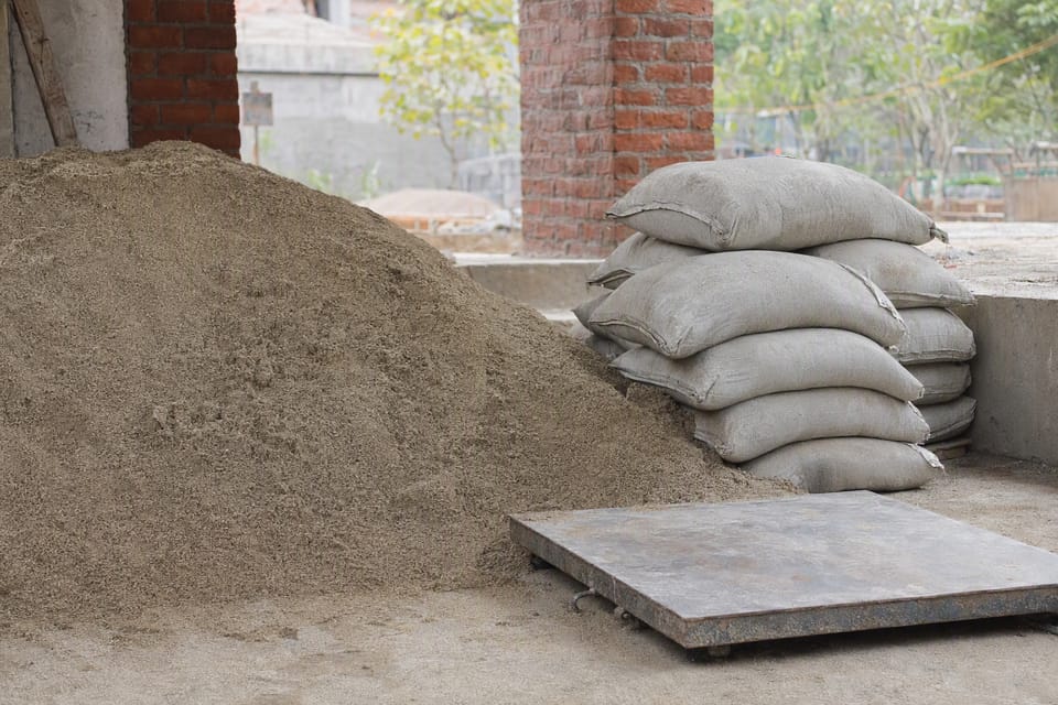 A heap of sand beside stacked cement bags and a steel mixing platform on a building site