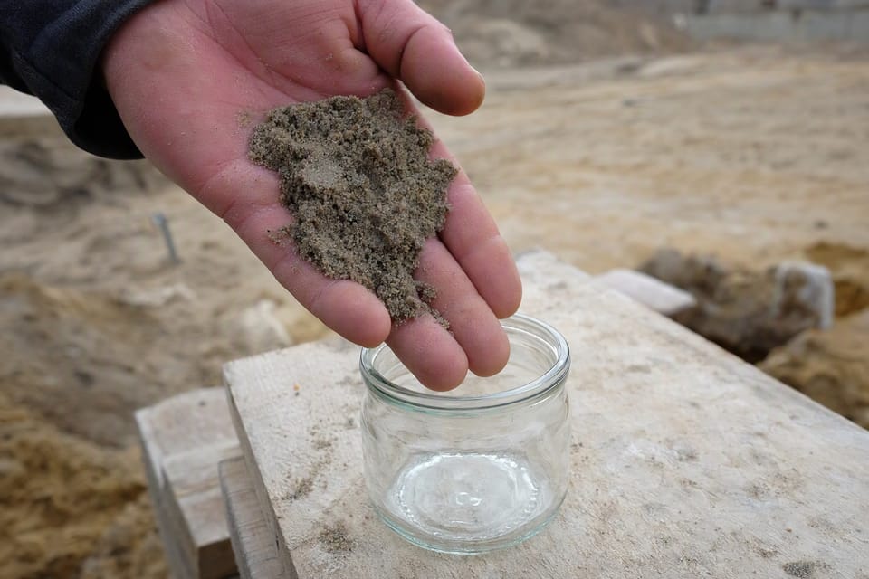 A hand holding a small amount of damp sand over a clear glass jar