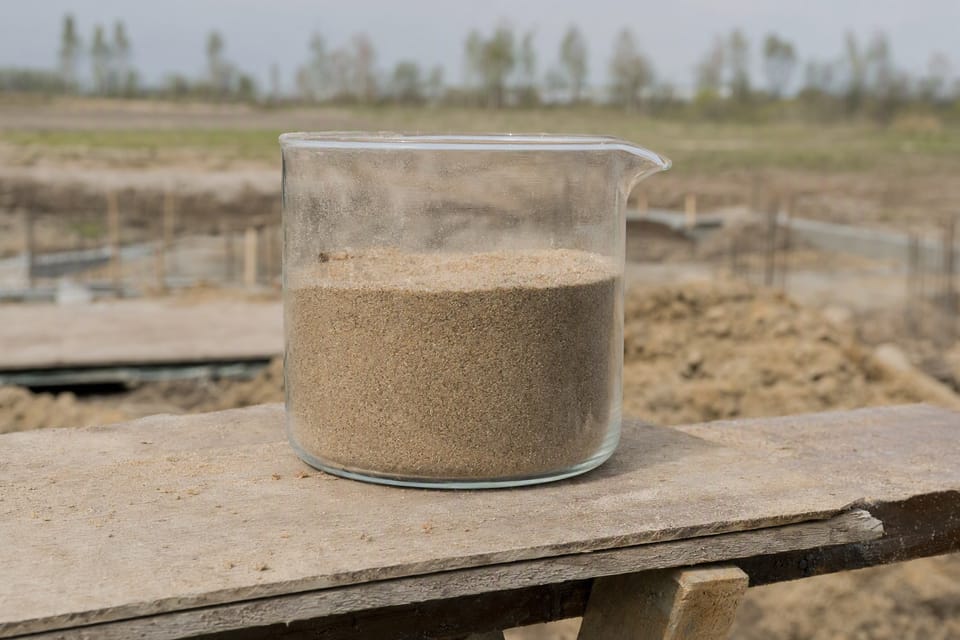A clear glass beaker half filled with sand standing on a wooden board