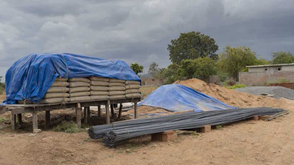 Cement bags on a raised platform under a tarpaulin, steel bars on bricks and a covered sand pile on a site under dark clouds