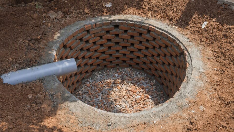A round soak pit being built in a yard, lined with bricks laid with open gaps, with stones at the bottom and an inlet pipe