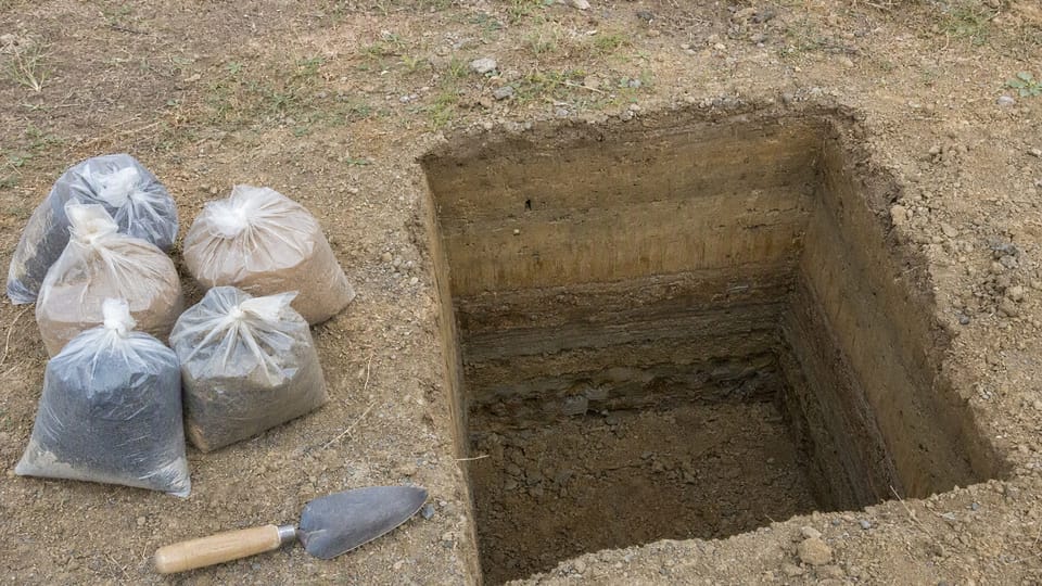 A square trial pit on a plot showing layers of soil, with sealed soil sample bags and a trowel beside it