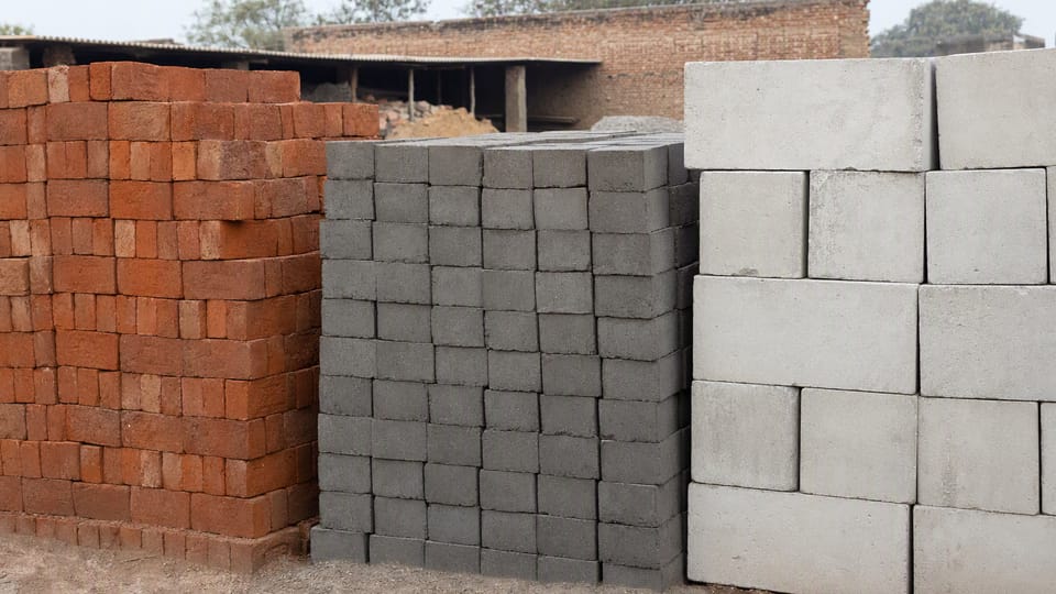 Three neat stacks of red clay bricks, grey bricks and larger pale blocks in a building-material yard