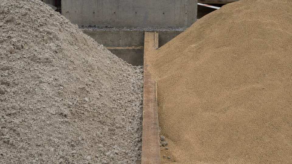 Two heaps of sand side by side on a building site, one pale and angular and one smoother and buff, with a wooden plank between them