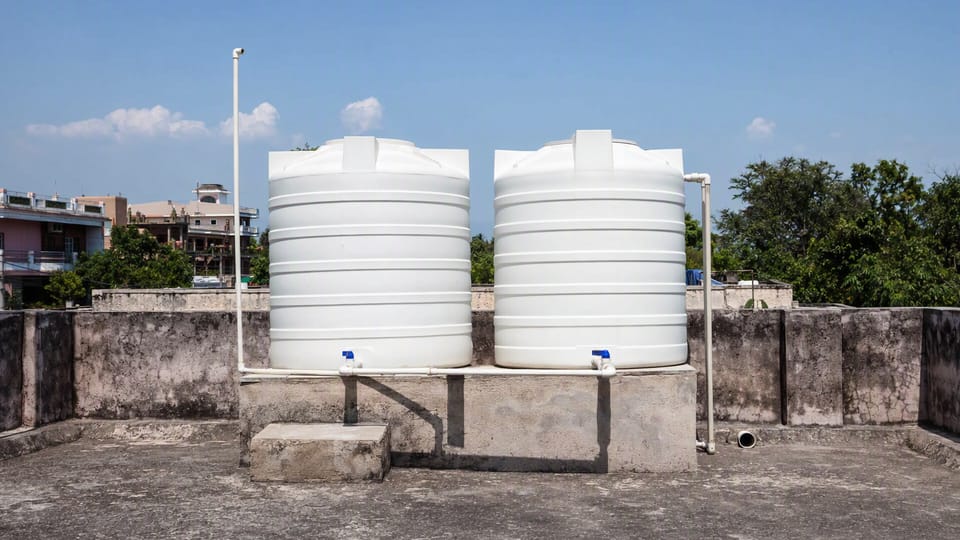 Two white cylindrical water tanks on a raised concrete platform on the terrace of an Indian house, with pipes and valves