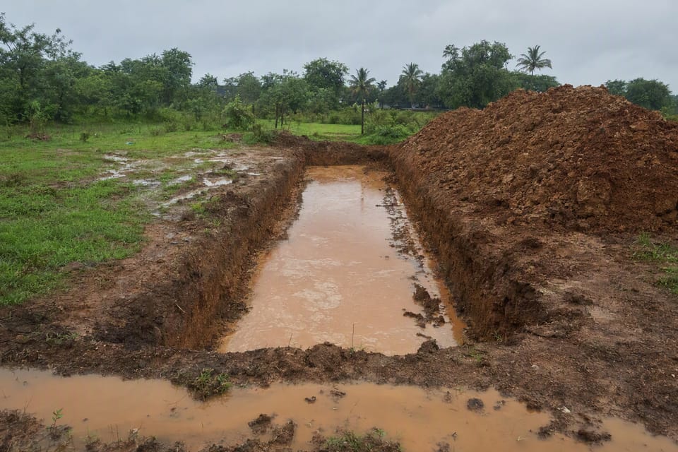 A rectangular foundation trench full of muddy rainwater beside a heap of excavated soil